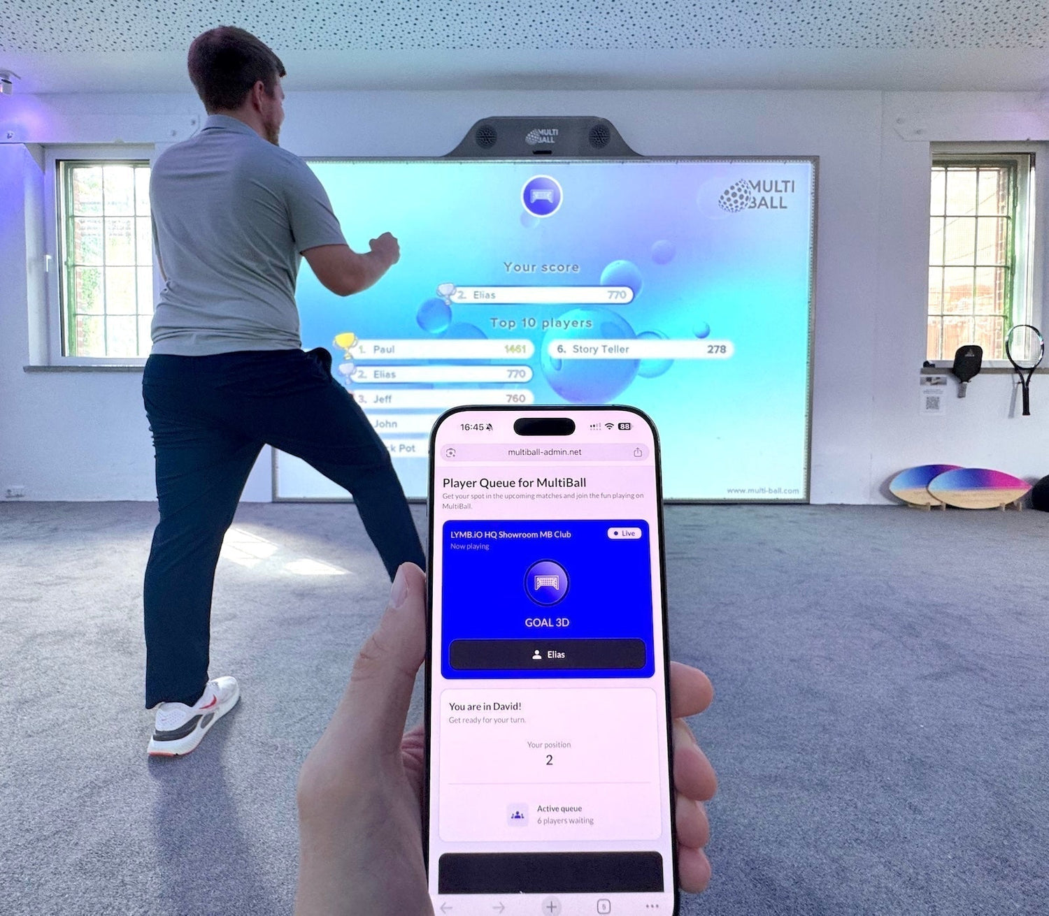 A smartphone showing the Player Queue for MultiBall with a player celebrating his score shown in the leaderboard on the interactive sports wall MultiBall in the background