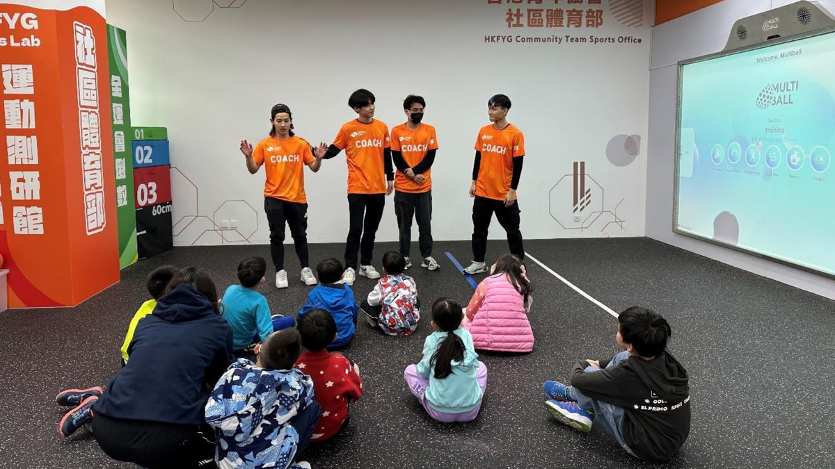 Four people in orange T-shirts teaching a group of little children how to use the interactive MultiBall system at the HKFYG Community Team Sports Office.