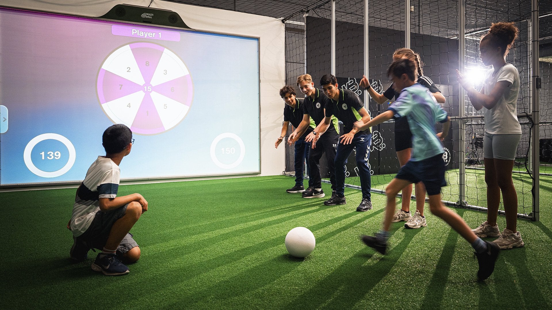 A child kicking a soccer ball at the sports simulation game Darts on the interactive screen MultiBall in the indoor sports facility Kickbase.nl while other children cheer him on