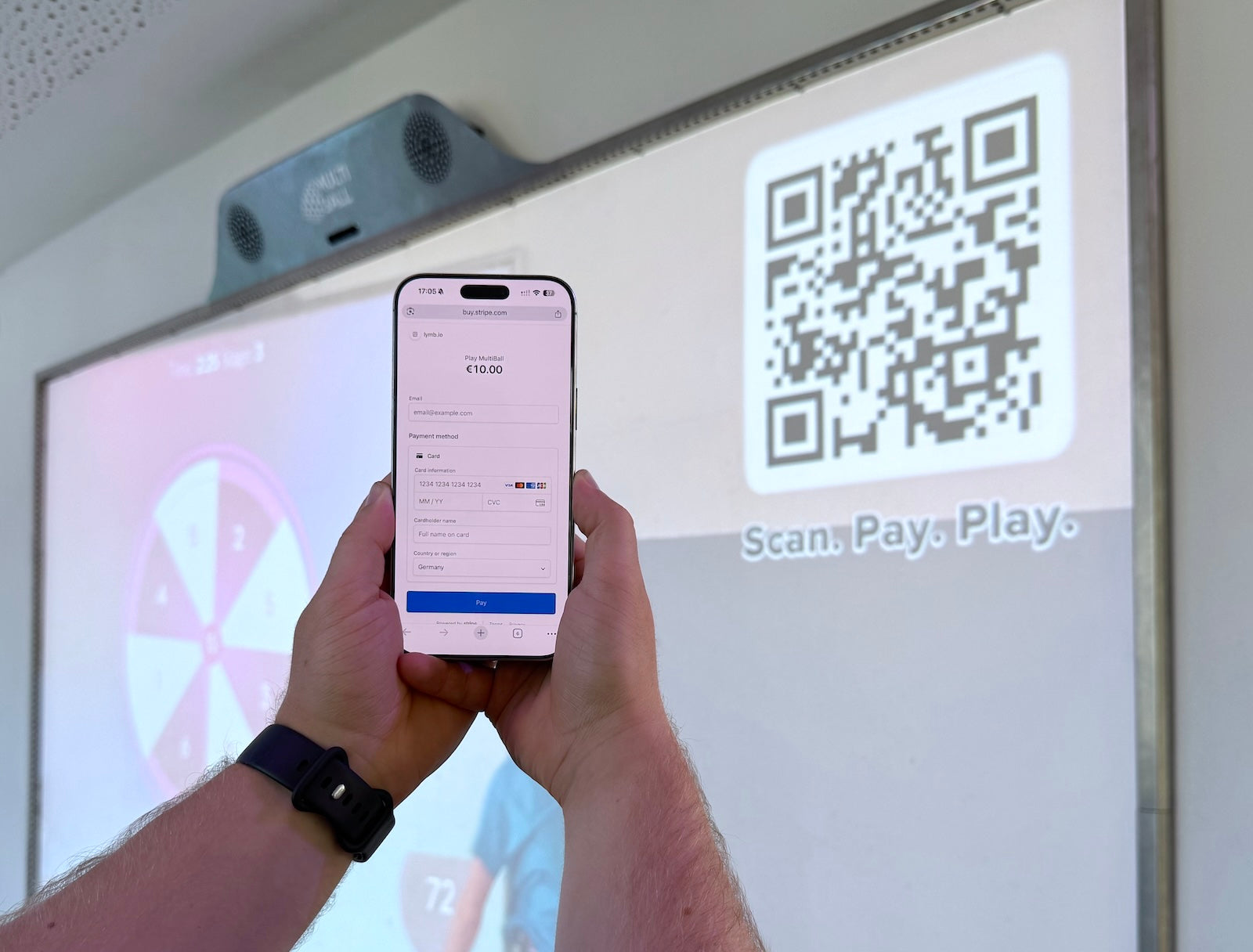 A person holding a smartphone with the Scan2Pay payment page, with the MultiBall in the background set to the Scan2Play mode and QR Code to pay per play time.