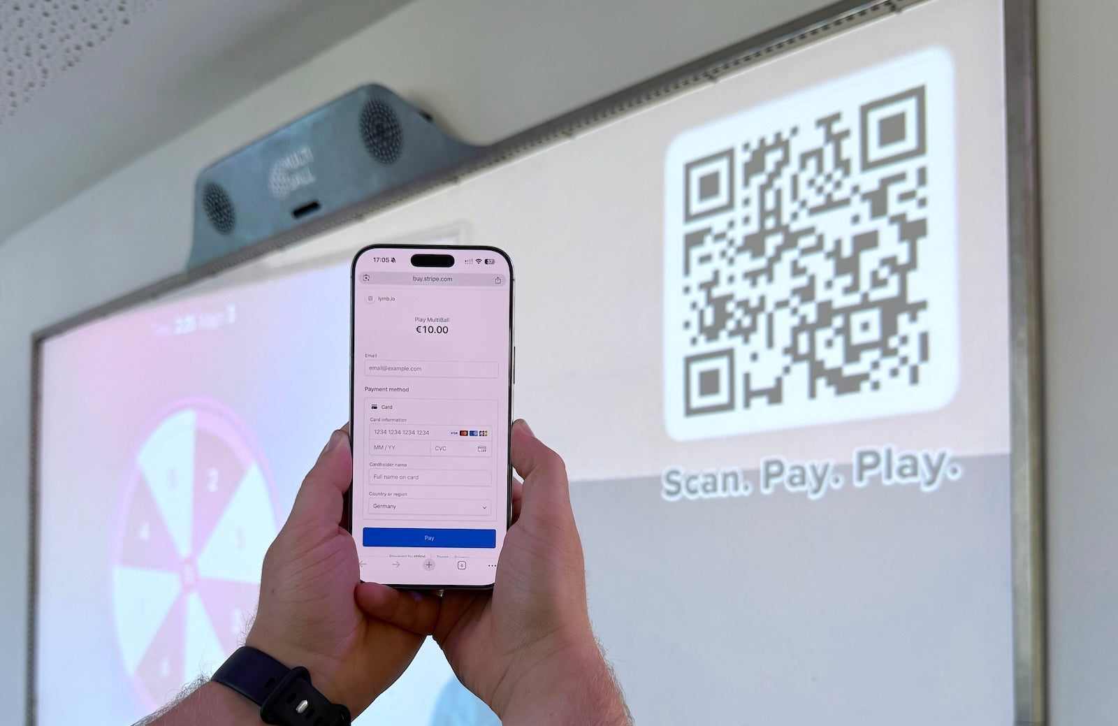 A person holding a smartphone with the Scan2Pay payment page, with the MultiBall in the background set to the Scan2Play mode and QR Code to pay per play time.
