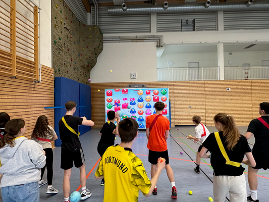 Students in a gymnasium engaging with a MultiBall interactive wall, throwing balls at targets on the screen during an educational game