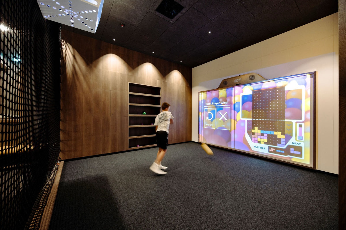 A kid playing the Block Drop game on the MultiBall in a nice room at Familien Resort Moar Gut, with a shelf in the back holding the balls, a net toon the left side and the projector with a projector cage installed on the ceiling.