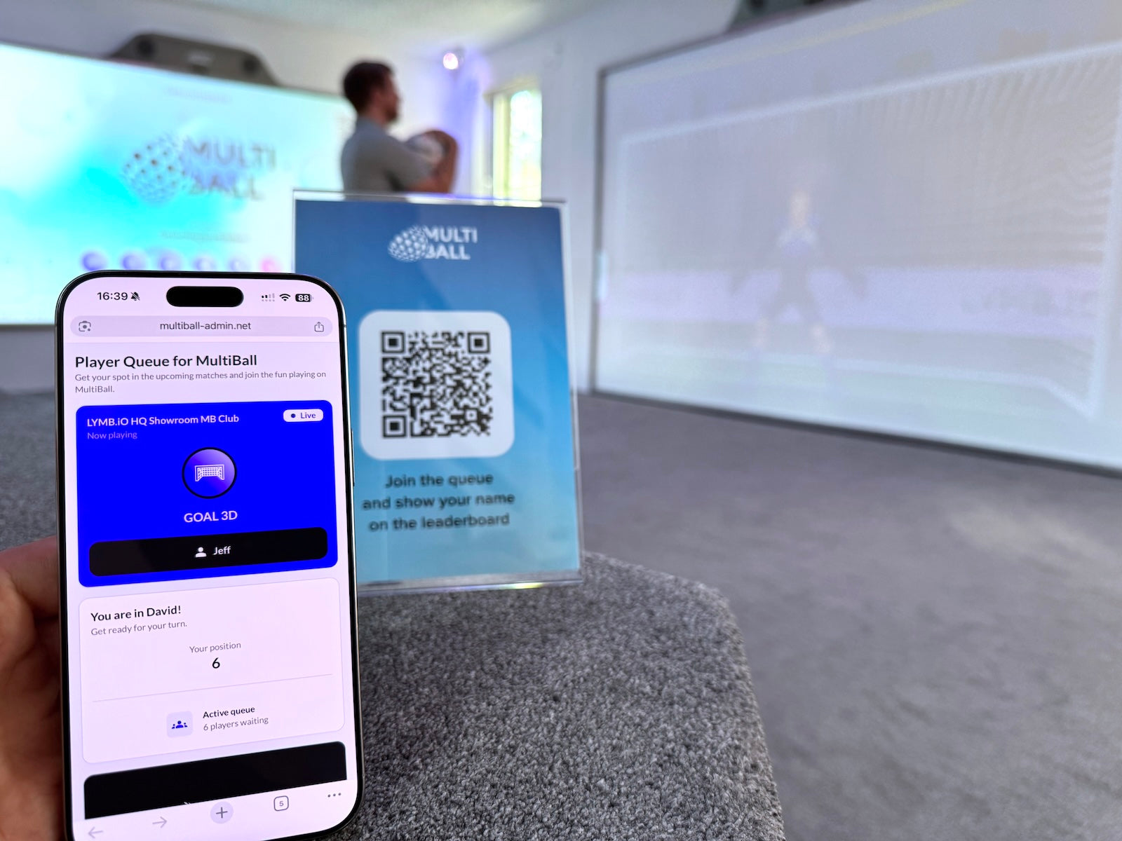 A Smartphone showing the that the person signed in is at position 6 in the Player Queue with the QR Code to join in the background and a person in front of a MultiBall system.