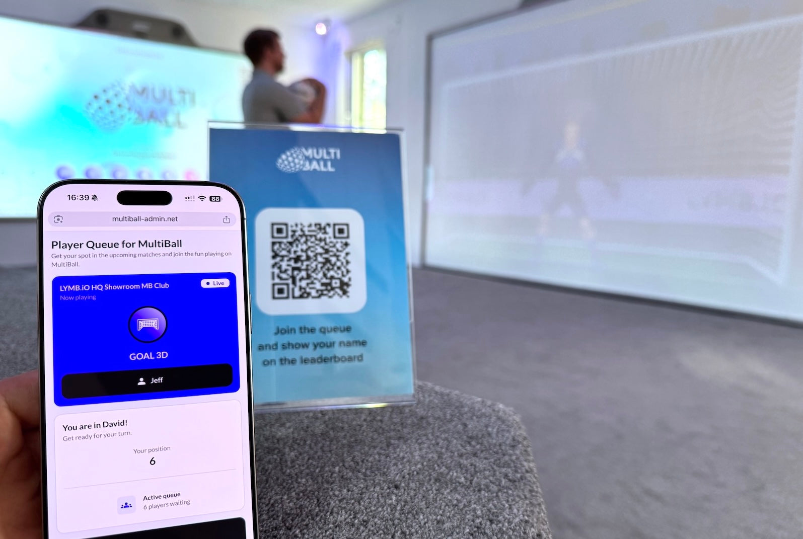 A Smartphone showing the that the person signed in is at position 6 in the Player Queue with the QR Code to join in the background and a person in front of a MultiBall system.