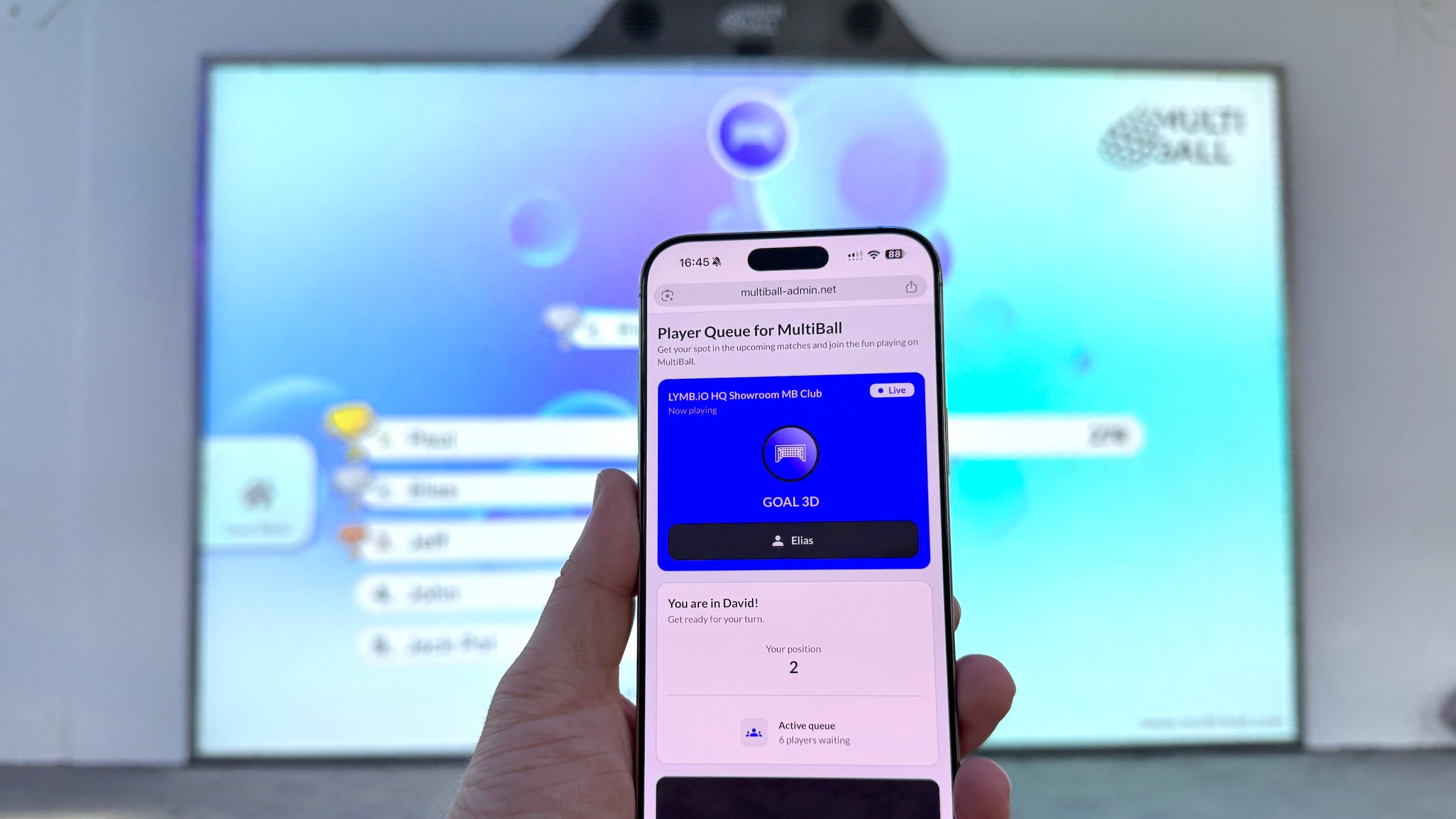A hand holds a smartphone displaying player view of the Player Queue open in the browser in the foreground, with the MultiBall showing the leaderboard in the background.