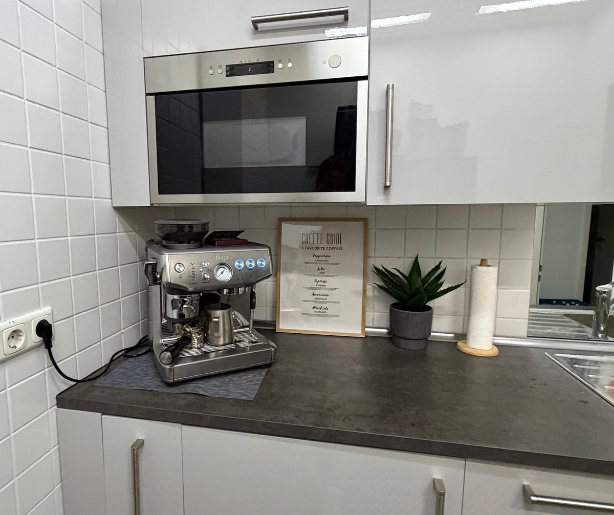A stainless steel espresso machine sits on a dark countertop in a modern kitchen setting with white cabinets, a tiled backsplash, and a small potted plant nearby as part of the PlayRooms at Frankfurter Ring