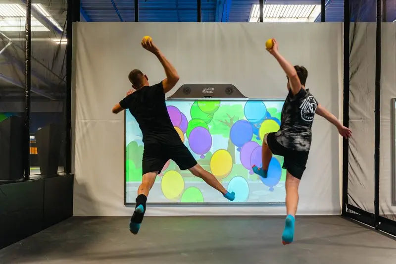 Two adults throwing balls while jumping on the interactive sports wall MultiBall in a trampoline park CyperJump.